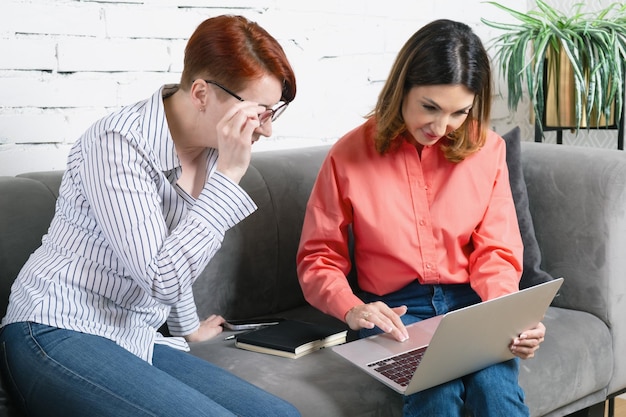 Zwei Geschäftsfrauen mittleren Alters sitzen auf einem Sofa im Büro und planen Arbeitsaufgaben. Frau mit schulterlangen Haaren zeigt Präsentation auf Laptop-Bildschirm. Treffen mit Kollegen