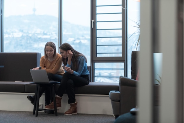 Zwei Geschäftsfrauen sitzen in einem modernen Coworking Space in einer Pause von der Arbeit und entspannen sich mit einem Laptop