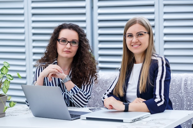 Zwei Geschäftsfrauen sitzen mit Laptop und Geschäftspapieren in einem hellen Café und unterhalten sich mit ernsthaften Leuten. Büroangestellte sitzen in Kaffeepausen und arbeiten. Geschäftstreffen in einem Café