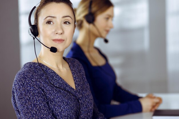 Zwei Geschäftsfrauen unterhalten sich mit den Kunden über Headsets, während sie in einem modernen Büro am Schreibtisch sitzen. Verschiedene Menschen gruppieren sich in einem Call Center. Telemarketing und Kundenservice.