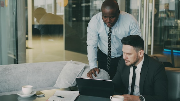 Zwei Geschäftspartner diskutieren aktiv ihr Startup-Projekt in einem stilvollen Café. Bärtiger Geschäftsmann sitzt und spricht mit seinem Kollegen, während er auf einen Laptop-Computer schaut