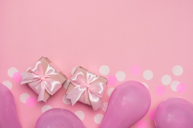 Zwei Geschenkboxen mit rosa Schleife auf pastellrosa Tisch mit weißen Konfetti und Luftballons