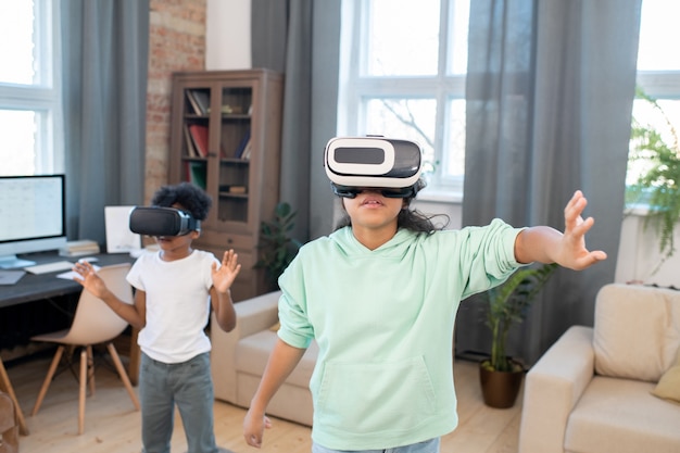 Zwei Geschwister in VR-Headsets und Freizeitkleidung schauen sich Online-Videos auf einem großen virtuellen Display an, während sie im Wohnzimmer vor der Kamera stehen