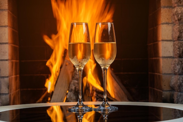 Zwei Gläser Champagner in der Nähe eines gemütlichen brennenden Kamins in einem Landhaus mit Reflexion an einer Glasoberfläche.