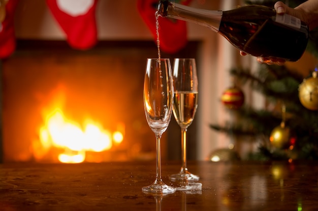 Zwei Gläser Champagner werden aus der Flasche gefüllt. Weihnachtsbaum und brennender Kamin im Hintergrund