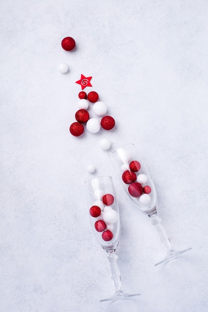 Zwei Gläser für Champagner und Weihnachts-Champagnerflasche mit Streuseln in Form eines Weihnachtsbaumes aus roten und weißen Spielzeugkugeln, dekoriert mit goldenem Konfetti auf Weiß.