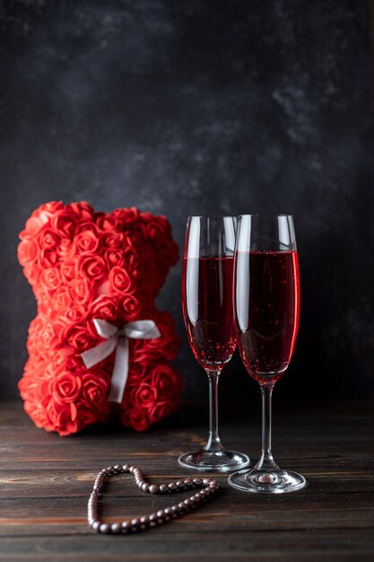 Zwei Gläser roter Champagner auf dunklem Hintergrund, ein Teddybär aus Rosen. Geschenk für Frauen. Frauenurlaub.