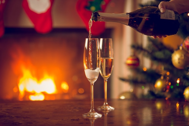 Zwei Gläser werden mit Champagner auf dem Tisch neben dem Weihnachtsbaum und dem brennenden Kamin gefüllt