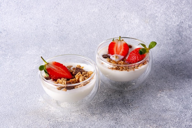 Zwei Glasbecher mit Müsli, Joghurt, Schokolade, Erdbeeren auf grauem Hintergrund