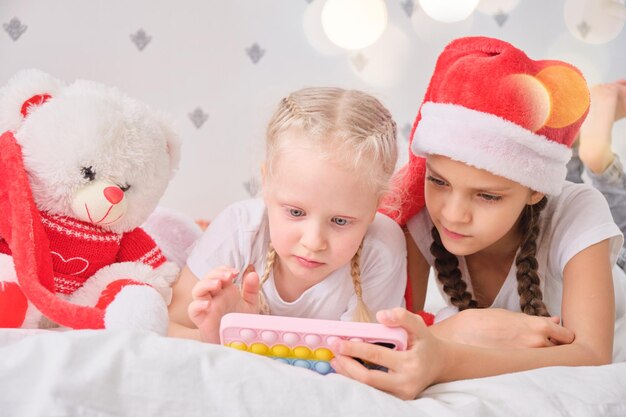 Zwei glücklich lächelnde Mädchen mit Smartphone auf dem Bett liegend Weihnachtsbeleuchtung Winterferien und Kinderkonzept, Kinder spielen Smartphone-Spiele, Weihnachtsstimmung