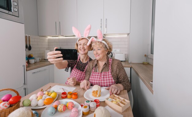 Zwei glückliche ältere Frauen mit Hasenohren auf dem Kopf fotografieren sich mit ihrem Handy, während sie sich zu Hause in der Küche auf Ostern vorbereiten.