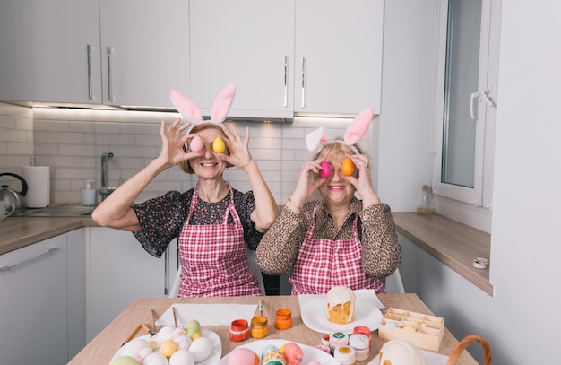Zwei glückliche ältere Frauen mit Hasenohren auf dem Kopf halten Ostereier an ihre Augen