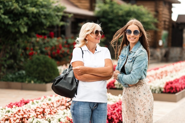 Zwei glückliche Frauen, ältere Mutter und lächelnde Tochter mit Sonnenbrille in modischem Denim-Outfit gehen in der Natur spazieren