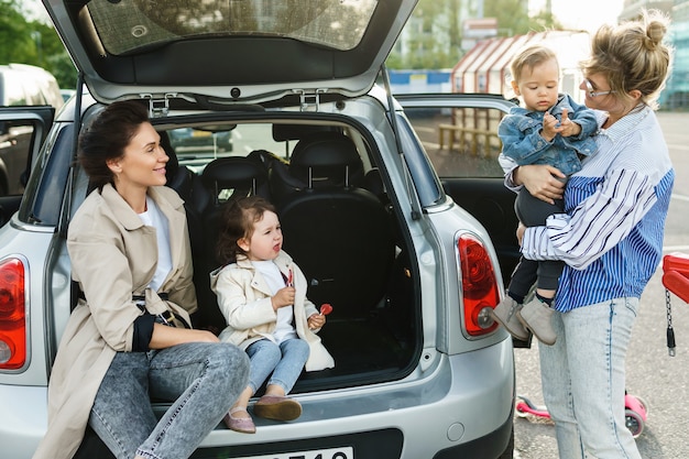 Zwei glückliche Frauen und ihre schönen Kinder neben einem Auto auf einem Parkplatz
