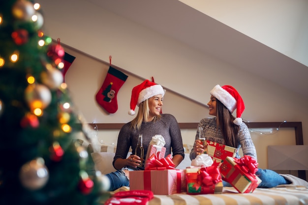 Zwei glückliche Freundinnen mit Weihnachtsmütze sitzen auf dem Bett für Weihnachtsferien und trinken Bär zufrieden