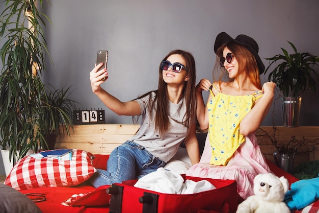 Zwei glückliche, fröhliche Freundinnen tragen eine Sonnenbrille und machen ein Selfie, das mit Kleidung und Koffer auf dem Bett sitzt