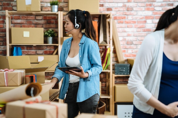 Zwei glückliche, fröhliche, schöne junge Telefonisten mit Headset, die in einem Online-Shop-Unternehmen arbeiten, das Verpackungsprodukte vorbereitet. asiatische kollegin mit schwangerer dame im büro