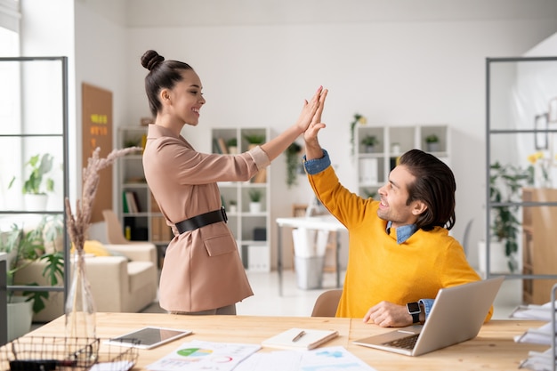 Zwei glückliche junge erfolgreiche Kollegen geben sich gegenseitig High Five, nachdem sie die Arbeit an einem neuen Projekt in einer Büroumgebung abgeschlossen haben