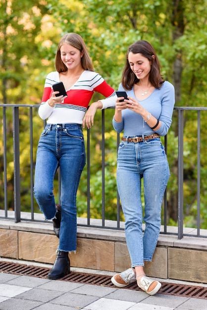 Zwei glückliche junge Frauen, die Seite an Seite gegen ein Geländer stehen, ihr Handy benutzen, das in ihre sozialen Medien vertieft ist und sich in einem Sucht- und Kommunikationskonzept ignorieren