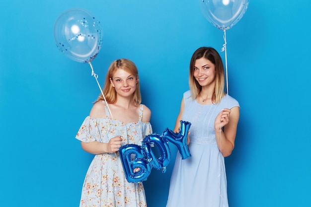 Zwei glückliche junge Frauen posieren in einem Studio mit blauen Luftballons und Ballon mit dem blauen Hintergrund des Aufschriftjungen