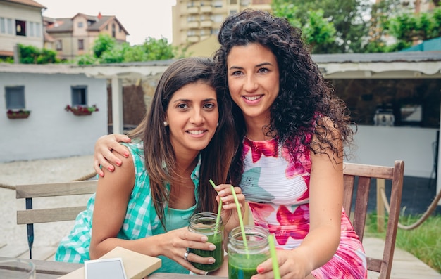 Zwei glückliche junge Frauen umarmten das Schauen, während sie grüne Gemüse-Smoothies im Freien hielten