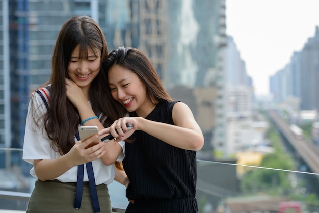 Zwei glückliche junge schöne asiatische Teenagerfrauen, die Telefon zusammen gegen Ansicht der Stadt verwenden