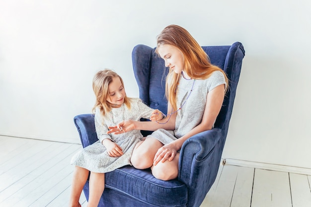 Zwei glückliche Kinder sitzen auf einem gemütlichen blauen Stuhl und spielen im weißen Wohnzimmer drinnen. Kleines Mädchen, das mit Teenager-Mädchen spielt, das ihre Liebessorge zeigt. Schwestern, die zu Hause Spaß haben.