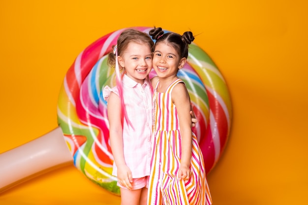 Zwei glückliche kleine Mädchen im bunten Kleid lachen umarmend und haben Spaß auf gelber Oberfläche mit Lutscher.