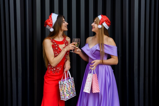 Zwei glückliche lächelnde Mädchen in den Weihnachtshüten, die mit Champagnergläsern rösten. Neujahrs- und Weihnachtsfeier