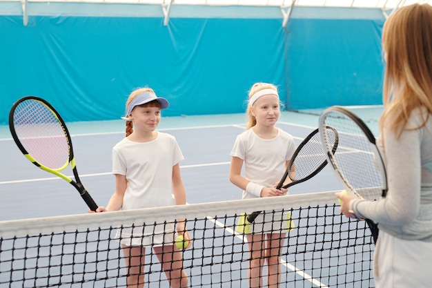 Zwei glückliche Mädchen in weißer Sportkleidung, die Tennisschläger halten, während sie vor ihrem Lehrer am Netz stehen und ihren Erklärungen zuhören