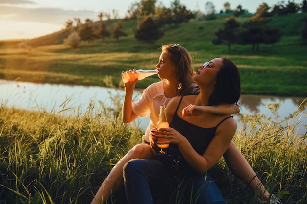 Zwei glückliche Mädchen trinken aus Flaschen, bei Sonnenuntergang, positivem Gesichtsausdruck, Outdoor-, Urlaubs- und Glückskonzept sind sie unten auf dem Rasen