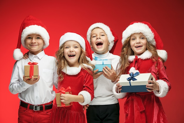 Zwei glückliche Mädchen und Jungen in Weihnachtsmannmützen mit Geschenkboxen im roten Studio