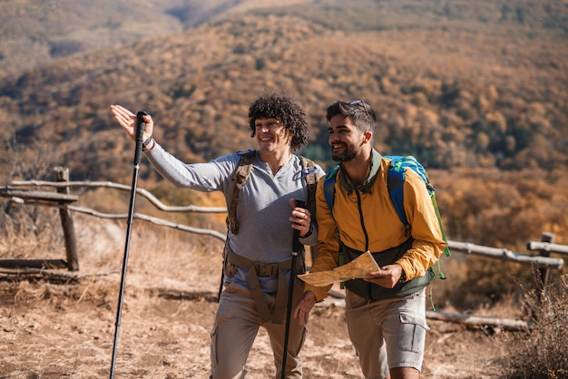 Zwei glückliche männliche Freund, die im Herbst wandern. Einer von ihnen hält die Karte, während der andere den richtigen Weg zeigt. Rucksäcke auf dem Rücken.