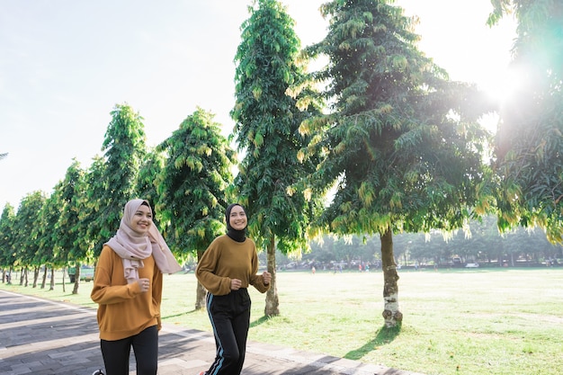 Zwei glückliche muslimische Mädchen mit Kopftüchern treiben Sport im Freien, während sie zusammen im Park mit Exemplar joggen