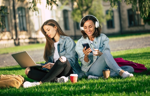 Zwei glückliche schöne junge Studentinnen in lässiger Jeanskleidung entspannen sich im College-Park mit Laptop und Smartphone von der Universität und trinken Kaffee.