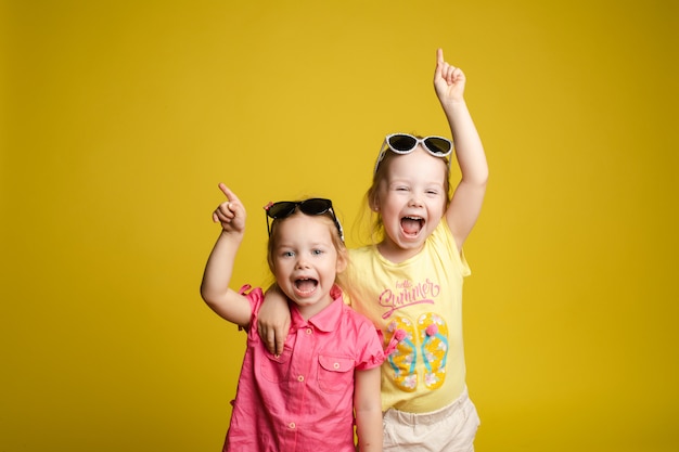 Zwei glückliche schöne stilvolle kleine Mädchen tragen Sonnenbrille posiert isoliert gelben Studio Hintergrund