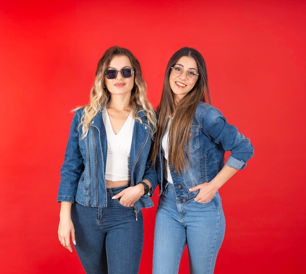 Zwei glückliche, stilvolle, modische Freundinnen posieren isoliert zusammen vor rotem Studiohintergrund