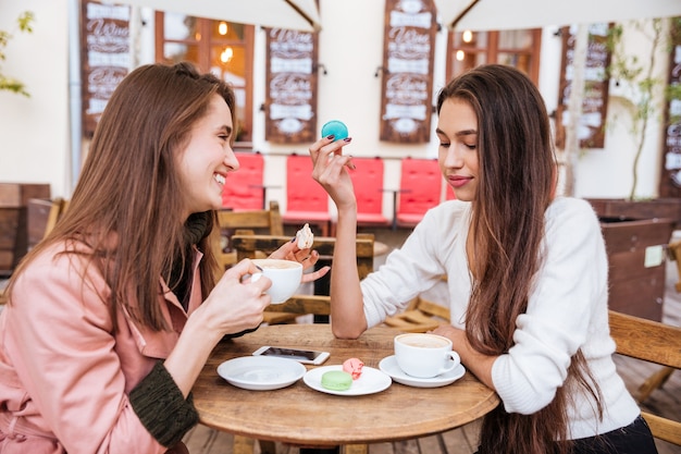 Zwei glückliche süße junge Frauen trinken Kaffee und essen französische Makronen im Café im Freien