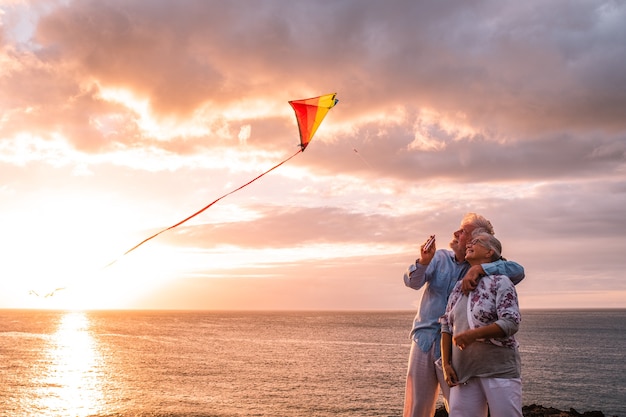 Zwei glückliche und fröhliche Senioren, die Spaß haben, einen fliegenden Drachen am Strand mit dem Sonnenuntergang und dem Meer im Hintergrund zu genießen und zu spielen - aktive Lifestyle-Menschen im Freien