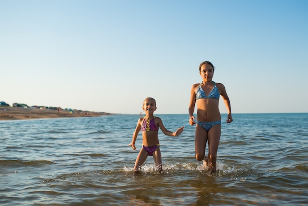 Zwei glückliche und positive Mädchenschwestern laufen während ihres Urlaubs an einem sonnigen heißen Sommertag entlang der Meereswellen