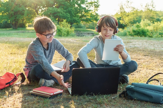 Zwei Grundschüler sitzen im Park auf dem Boden und lernen mit einem Laptop. Junge mit Brille hält Arbeitsbuch auf dem Schoß und zeigt auf ein Gerät auf einen Schüler mit Kopfhörern, der eine Aufgabe im Notizbuch liest