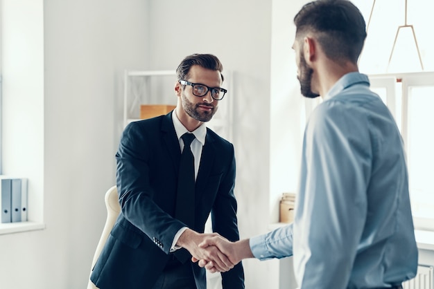 Zwei gut aussehende junge Männer in formeller Kleidung, die sich im Büro die Hände schütteln