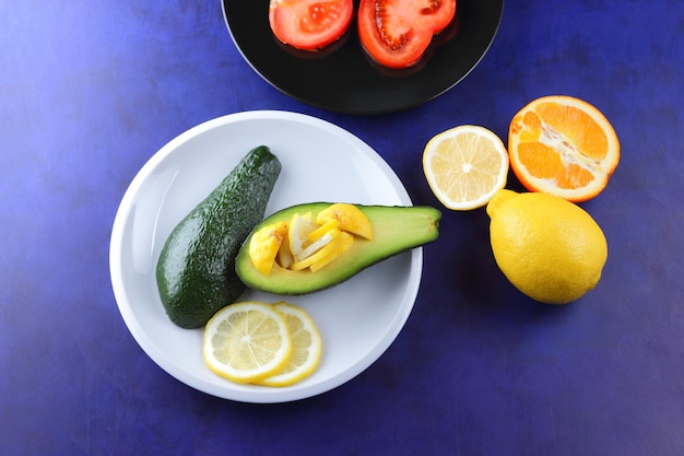 Zwei Hälften einer reifen Avocado mit Samen auf einem Teller Nahaufnahme von grünen Früchten und Zitrus- und Tomatenscheiben auf blauem Hintergrund Vegetarisches Ernährungskonzept