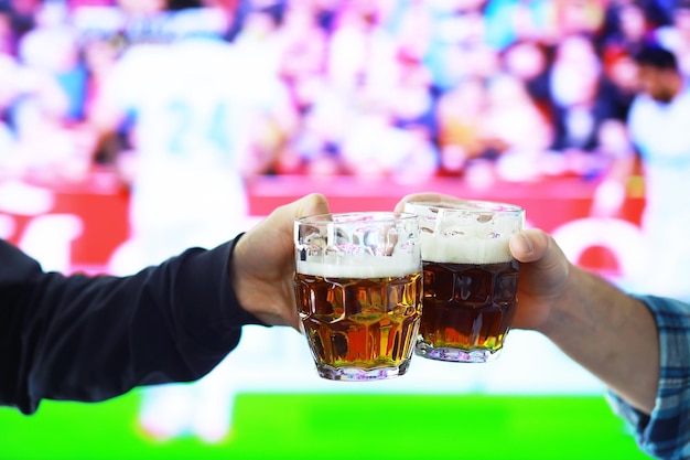 Zwei Hände, die horizontales Lagerbierglas halten und auf dem Hintergrund des Fußballspiels klirren. Sportfans jubeln. Freunde Freizeit-Lifestyle-Konzept.