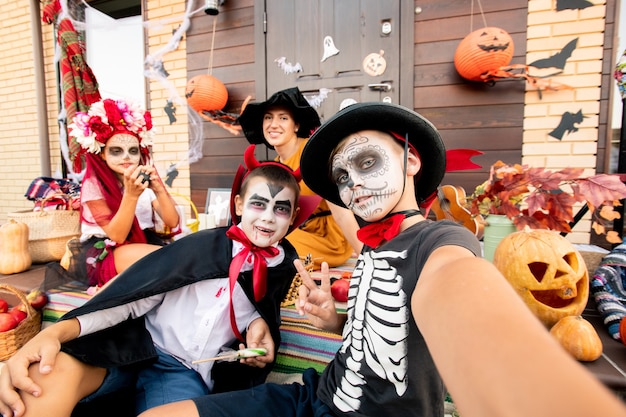 Zwei Halloween-Jungen machen Selfie beim Sitzen auf der Treppe auf dem Hintergrund eines süßen Mädchens und einer glücklichen jungen Frau in Hexenhut und gelbem Kleid