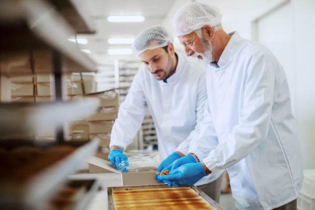 Zwei hart arbeitende Mitarbeiter der Lebensmittelfabrik in sterilen Uniformen, die Kekse in Kisten verpacken.