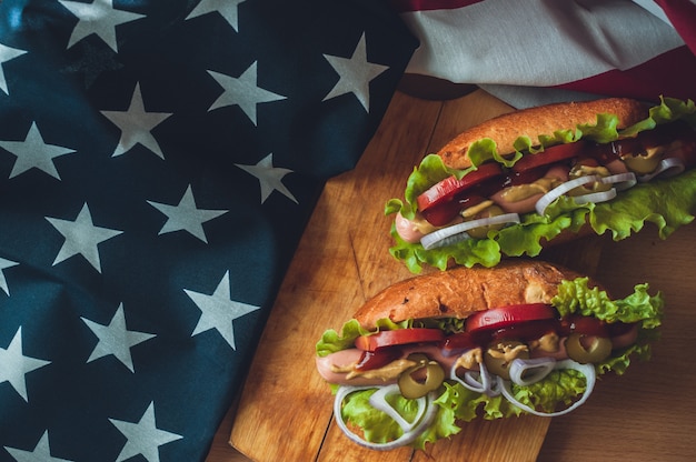 Zwei Hotdogs auf einem hölzernen Brett, Gläsern mit Kolabaum und amerikanischer Flagge