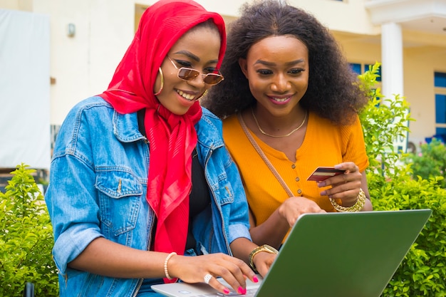 Zwei hübsche afrikanische Damen, die ihre Kreditkarte und ihren Laptop benutzen, um online einzukaufen.