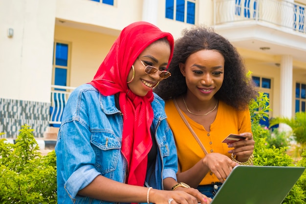 Zwei hübsche afrikanische Damen, die ihre Kreditkarte und ihren Laptop benutzen, um online einzukaufen.