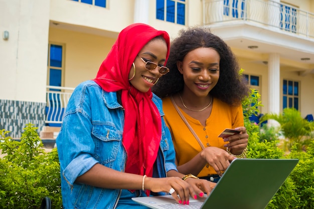 Zwei hübsche afrikanische Damen, die ihre Kreditkarte und ihren Laptop benutzen, um online einzukaufen.